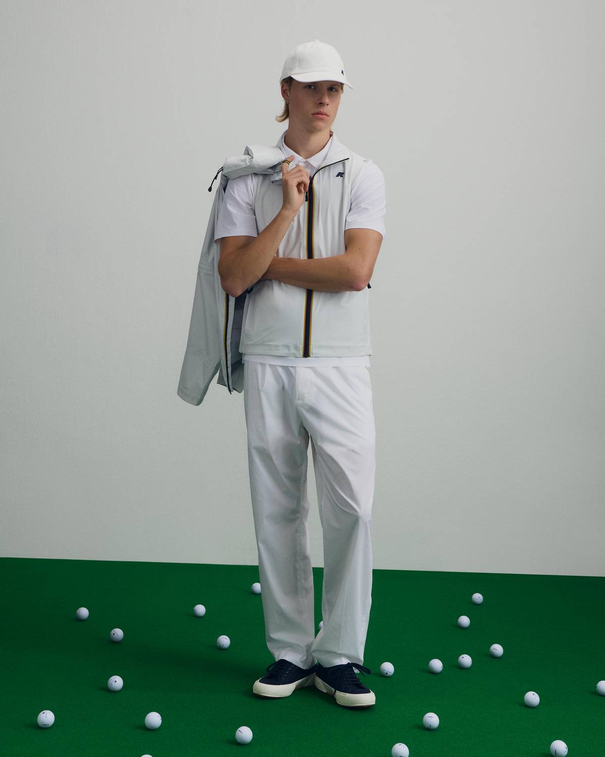 Golfista in completo bianco K-Way con gilet tecnico, cappellino, polo e sneakers blu su prato