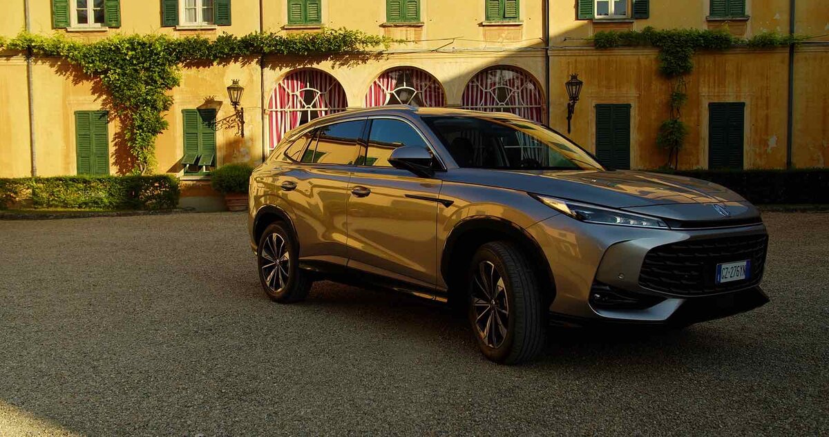 MG HS Hybrid+ grigia in vista tre quarti anteriore, parcheggiata in un cortile storico con edificio giallo sullo sfondo.