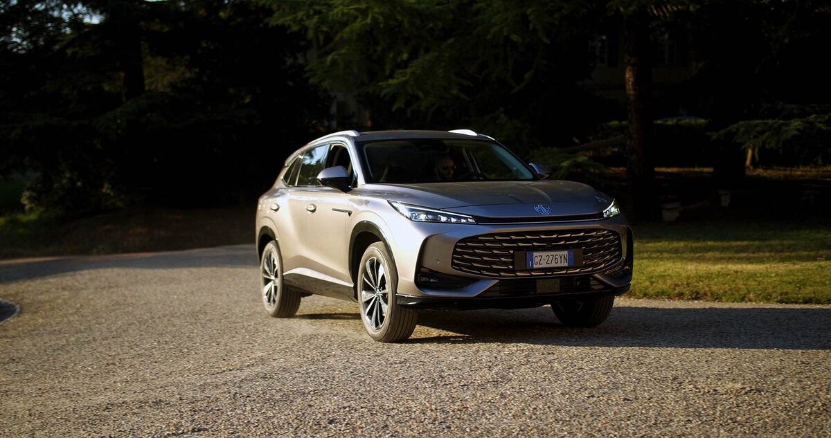 MG HS Hybrid+ grigia in movimento su strada sterrata, vista frontale in controluce con sfondo alberato.