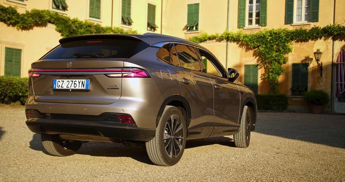 MG HS Hybrid+ grigia vista posteriore tre quarti, parcheggiata in un cortile con villa storica gialla e persiane verdi sullo sfondo.