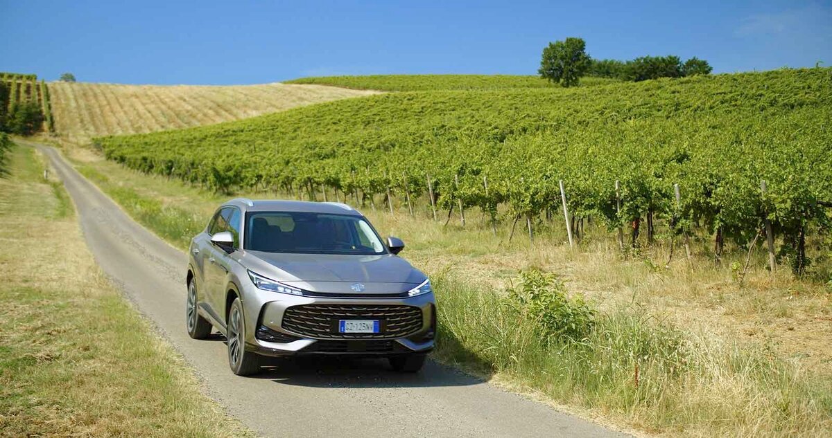 MG HS Hybrid+ grigia ripresa frontalmente mentre percorre una strada di campagna tra vigneti sotto cielo estivo limpido.