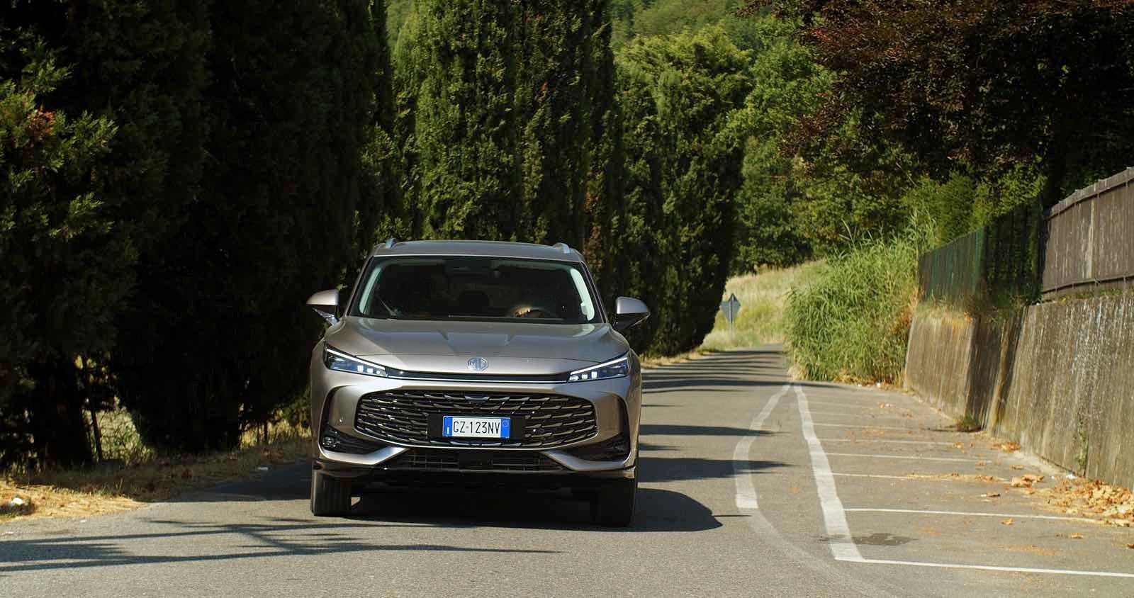 MG HS Hybrid+ grigia vista frontale su strada alberata di campagna, tra cipressi e vegetazione estiva in pieno giorno.