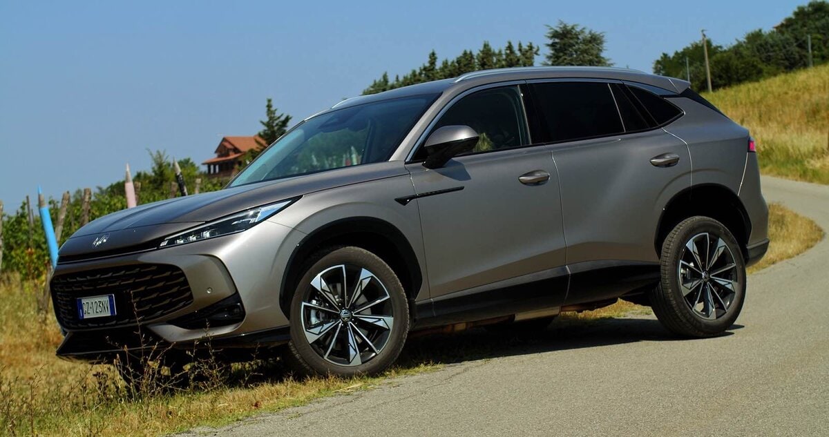 MG HS Hybrid+ grigia in vista laterale anteriore su strada di collina, con fondo asfaltato e vigneti in lontananza.