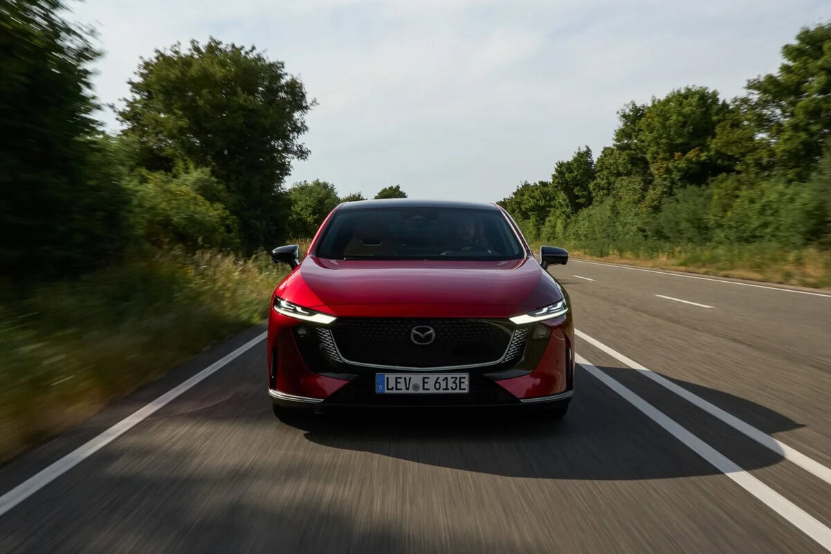 Frontale della Mazda 6e 2025 EV rossa che percorre una strada fiancheggiata da alberi.