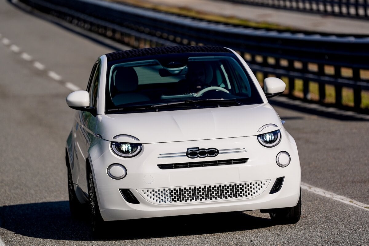 Nuova Fiat 500 ibrida bianca su strada durante i test, vista frontale con fari accesi e design compatto riconoscibile.