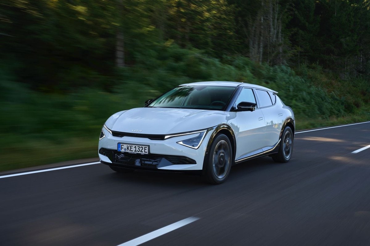 Kia EV6 bianca 2025 in movimento su strada asfaltata tra vegetazione e alberi in contesto naturale.