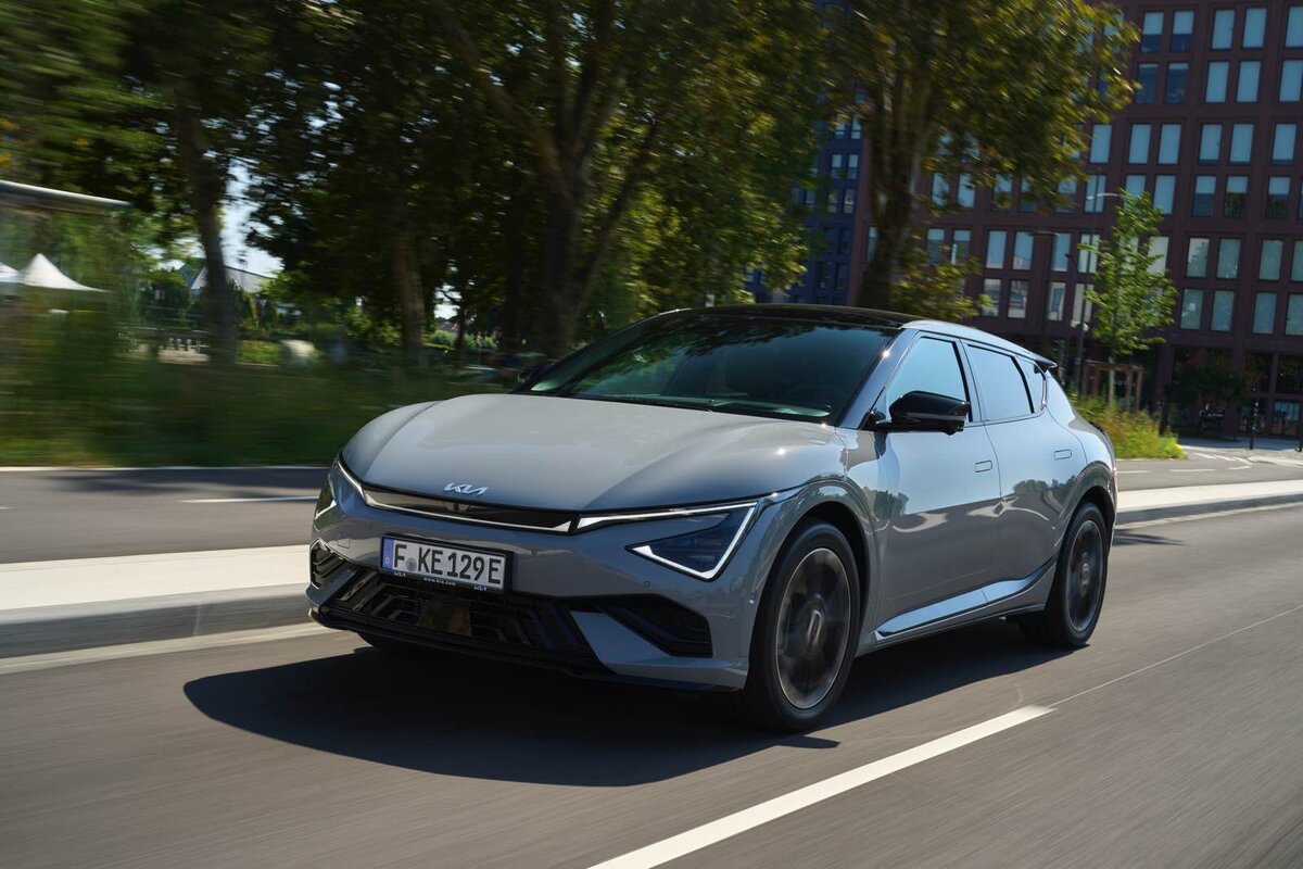 Kia EV6 grigia in corsa su strada cittadina con edifici moderni sullo sfondo, crossover elettrica dallo stile sportivo.