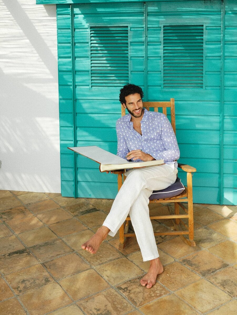 Uomo sorride su sedia in veranda, indossa camicia azzurra fantasia e pantaloni bianchi