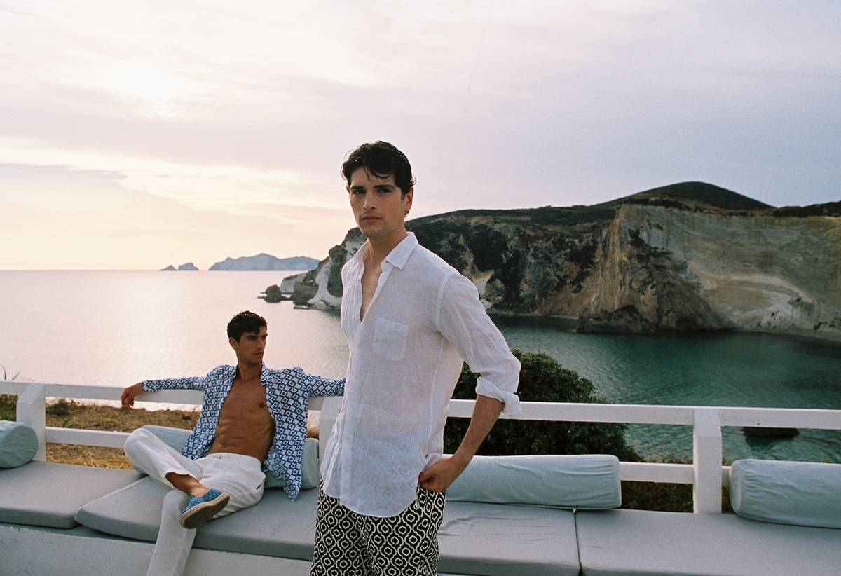 Due uomini con camicie Peninsula e pantaloni leggeri in terrazza al tramonto con vista mare