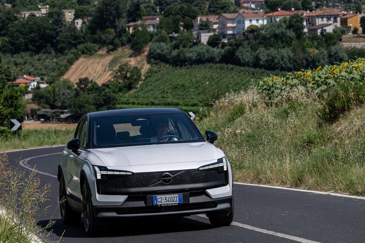 Volvo EX30 Cross Country MY26 in marcia su una strada collinare asfaltata, circondata da girasoli e vegetazione rurale.