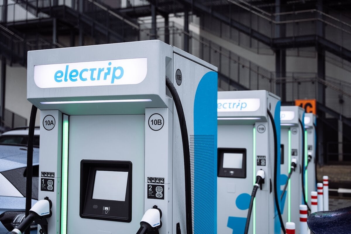 Stazioni di ricarica rapida per veicoli elettrici del marchio "electrip", con colonnine numerate e dotate di schermi e connettori CCS. Le unità sono disposte in fila in un ambiente urbano moderno, con dettagli architettonici in metallo sullo sfondo.