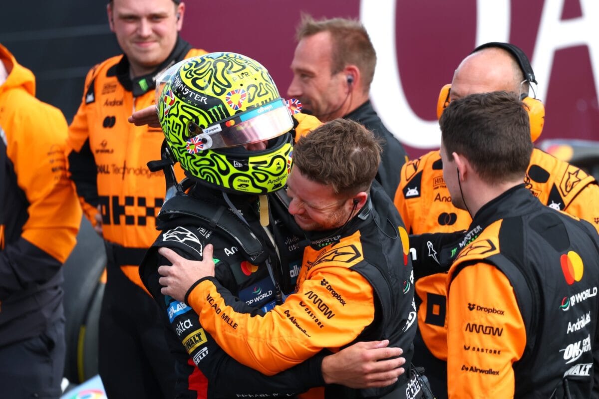 Lando Norris abbraccia un membro del team McLaren dopo la vittoria nel GP di Silverstone 2025, circondato dai meccanici in tuta arancione.
