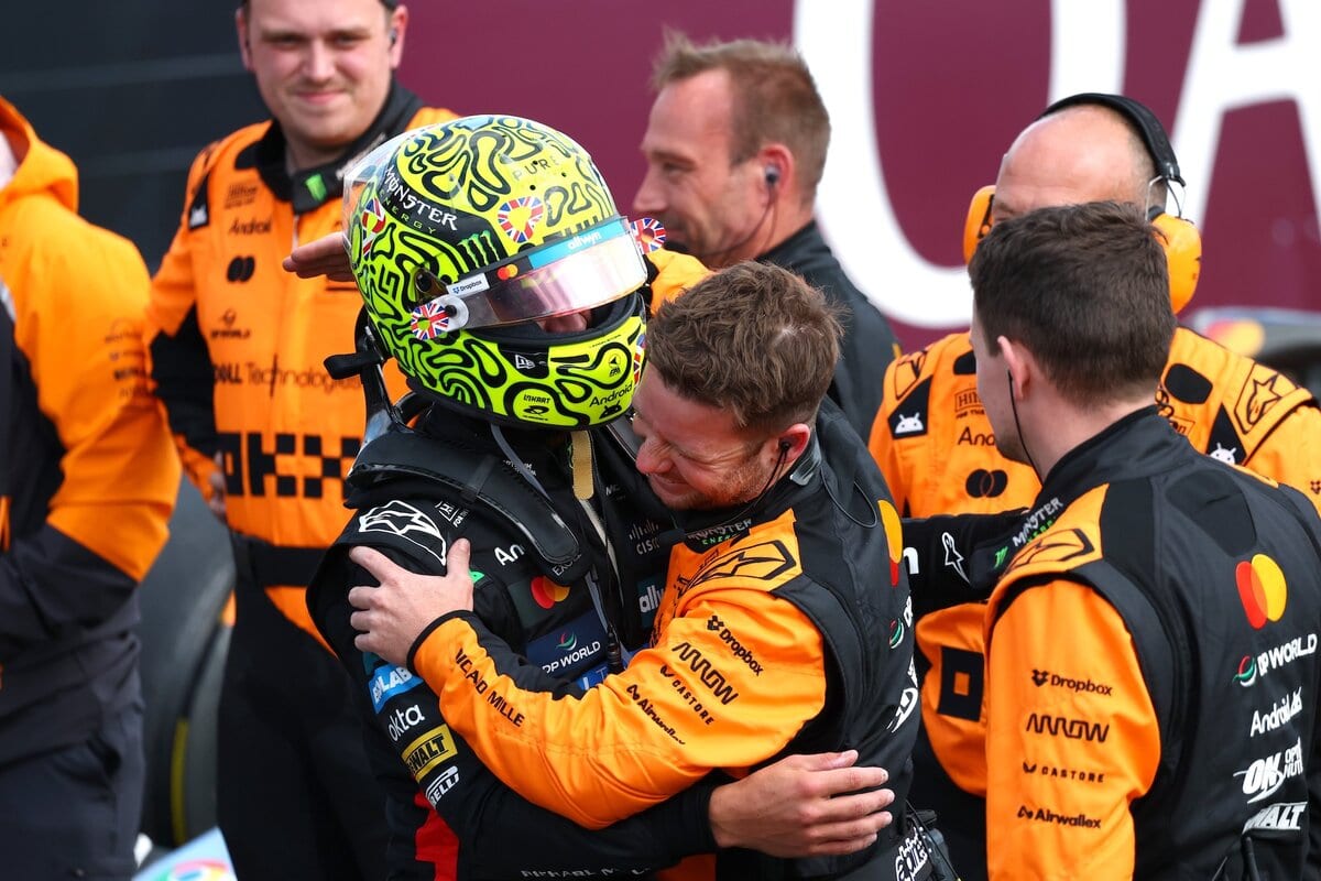 Lando Norris abbraccia un membro del team McLaren dopo la vittoria nel GP di Silverstone 2025, circondato dai meccanici in tuta arancione.