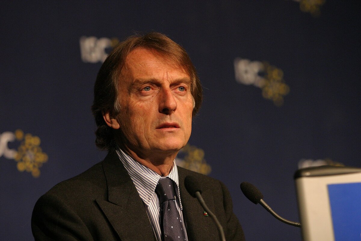 Luca Cordero di Montezemolo durante un intervento pubblico, in piedi davanti a un podio, con sfondo blu e microfono.