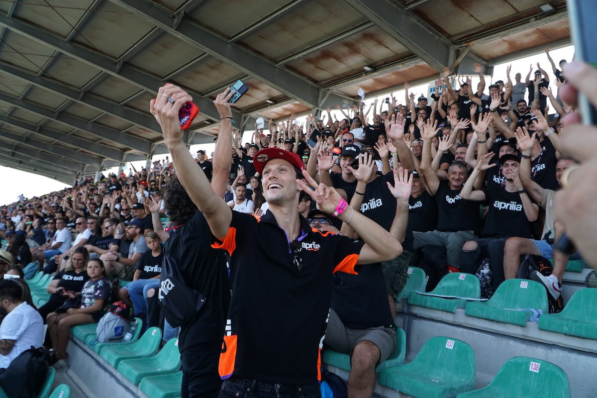 Fan della tribuna Aprilia al MotoGP di Misano posano sorridenti per un selfie di gruppo. Indossano magliette nere con logo Aprilia e sollevano le mani in segno di festa sotto la copertura della tribuna.