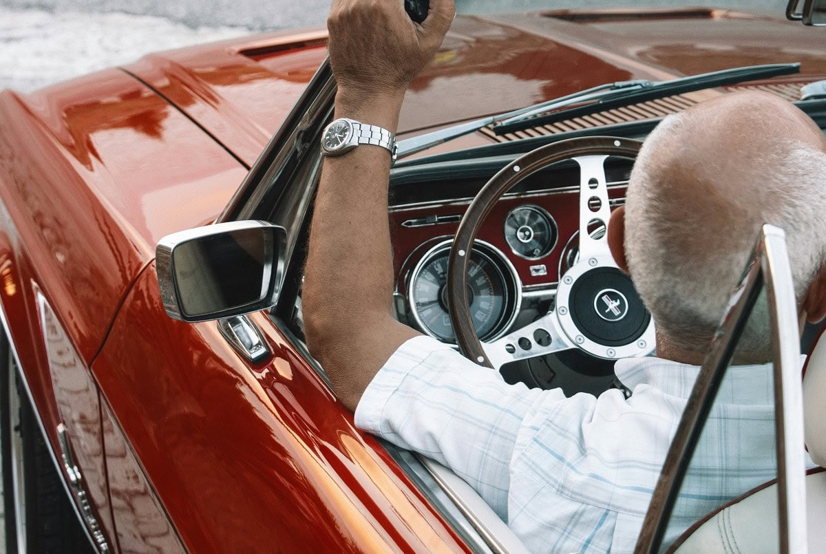 Un uomo anziano con la testa calva siede al posto di guida di una classica auto a noleggio, tenendo il volante con il braccio sinistro appoggiato alla portiera.