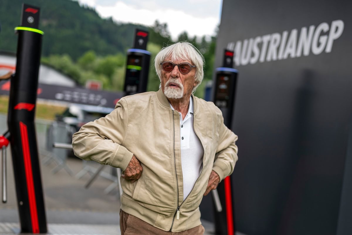 Bernie Ecclestone con capelli bianchi e occhiali da sole indossa una giacca beige e si sistema con le mani ai fianchi davanti