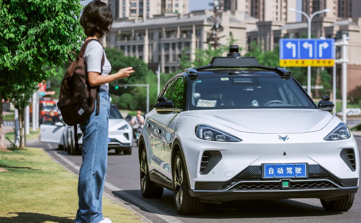 Una giovane donna con zaino si avvicina a un’auto a guida autonoma Pony.ai Apollo Go ferma sul marciapiede, con sensori Lidar sul tetto e targa blu cinese