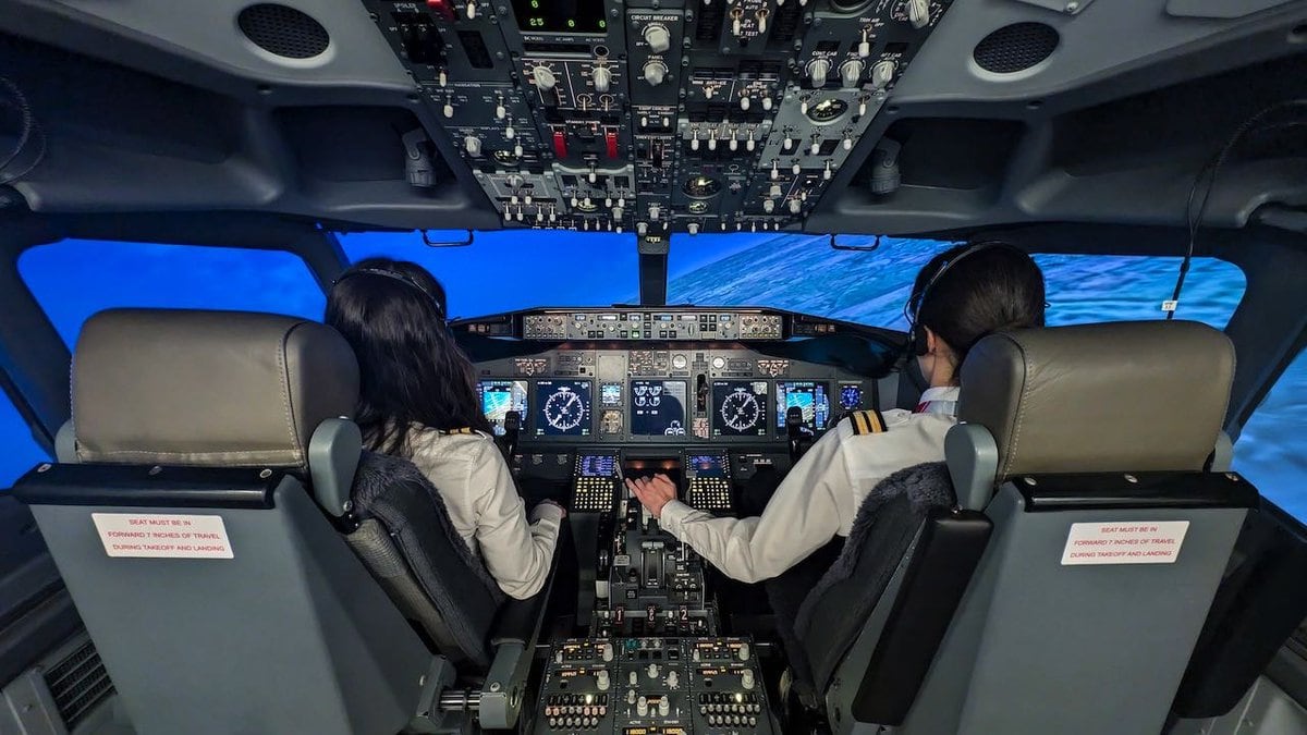 Due aspiranti piloti seduti nella cabina di un simulatore di volo, circondati da comandi realistici e schermi digitali, con vista virtuale sull'oceano attraverso i finestrini.
