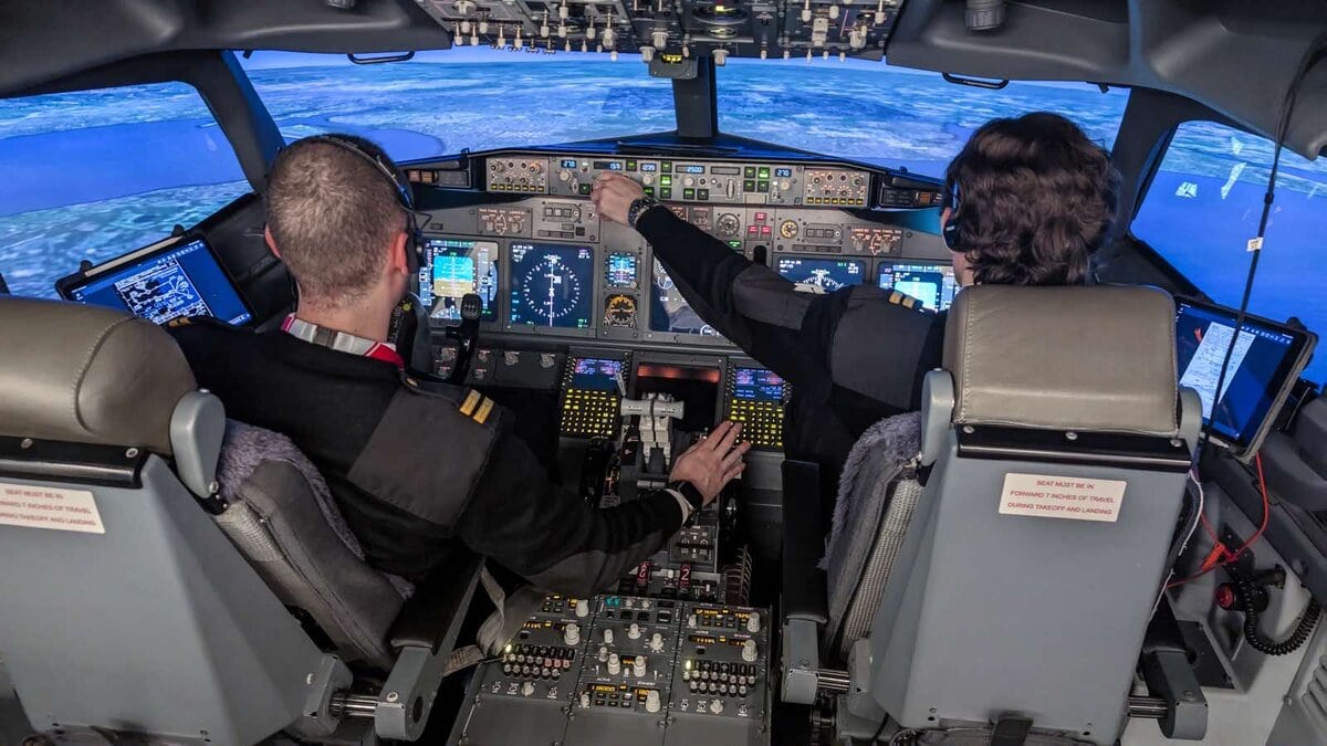 Due aspiranti piloti in divisa all’interno di un simulatore di volo avanzato, intenti a gestire i comandi del cockpit con il panorama digitale di una costa sullo sfondo.