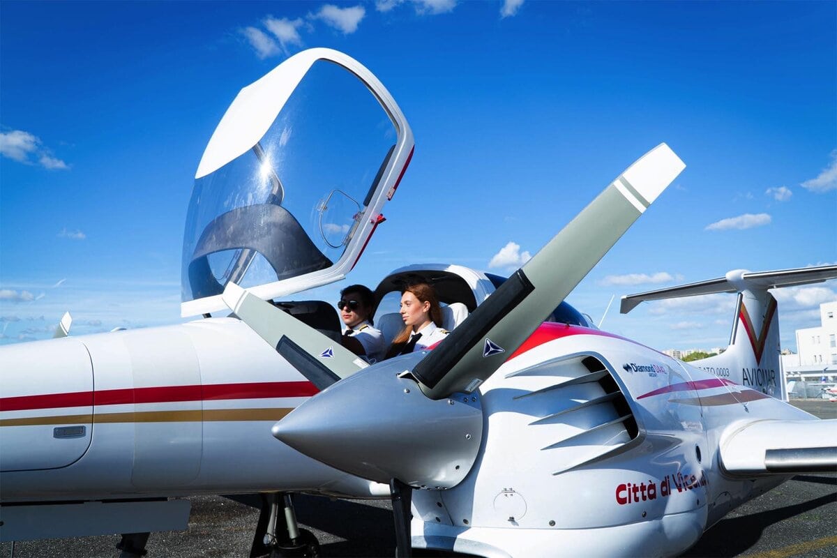 Due allievi piloti seduti nella cabina di un aereo da addestramento biposto a elica, con tettuccio aperto e cielo azzurro sullo sfondo.