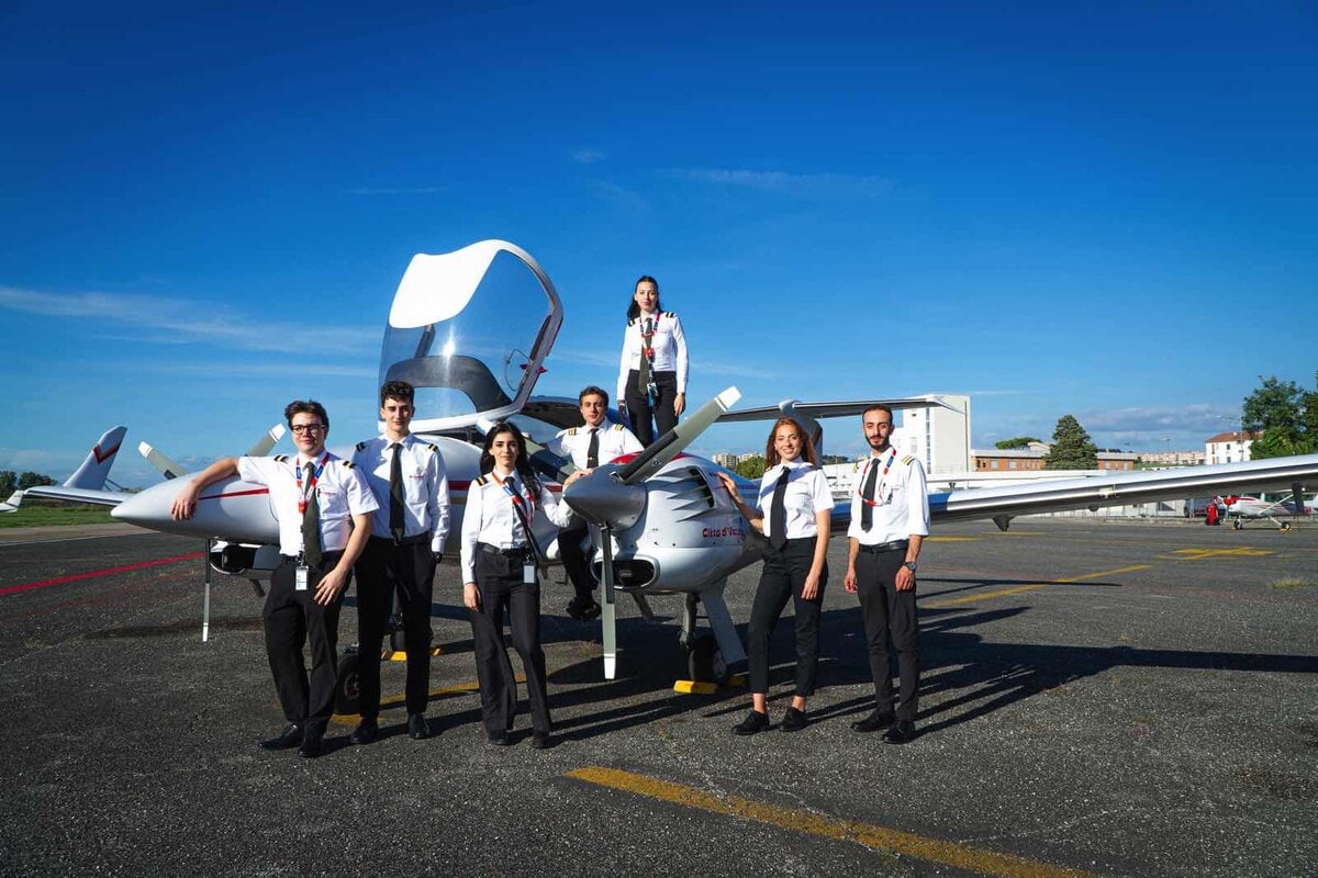 Gruppo di giovani cadetti piloti in uniforme schierati davanti a un aereo da addestramento Aviomar, con cielo azzurro e pista di volo sullo sfondo.