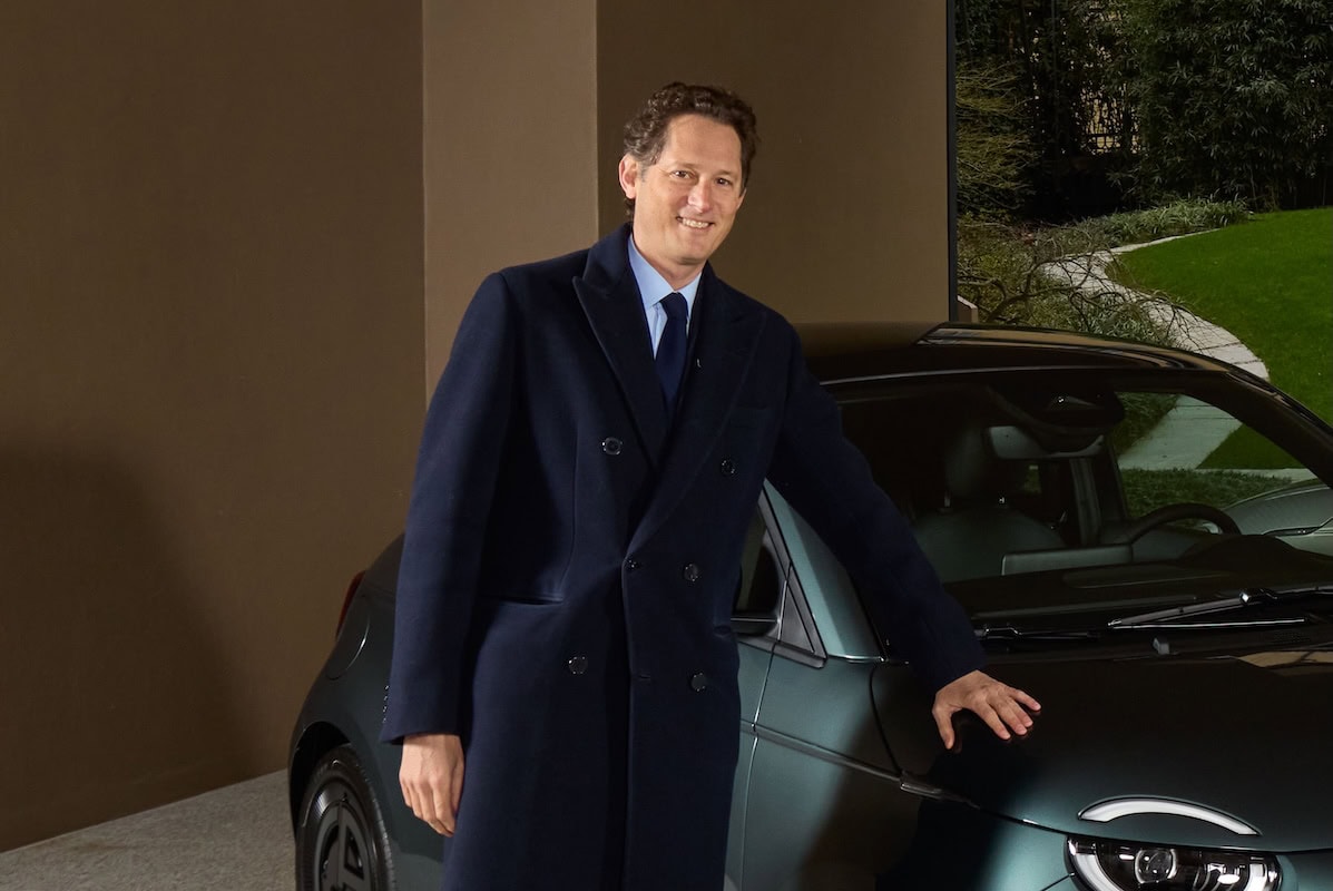 John Elkann in piedi accanto ad una Fiat 500 elettrica verde scuro, appoggiato sul cofano con una mano, indossa un cappotto blu elegante in un ambiente moderno.