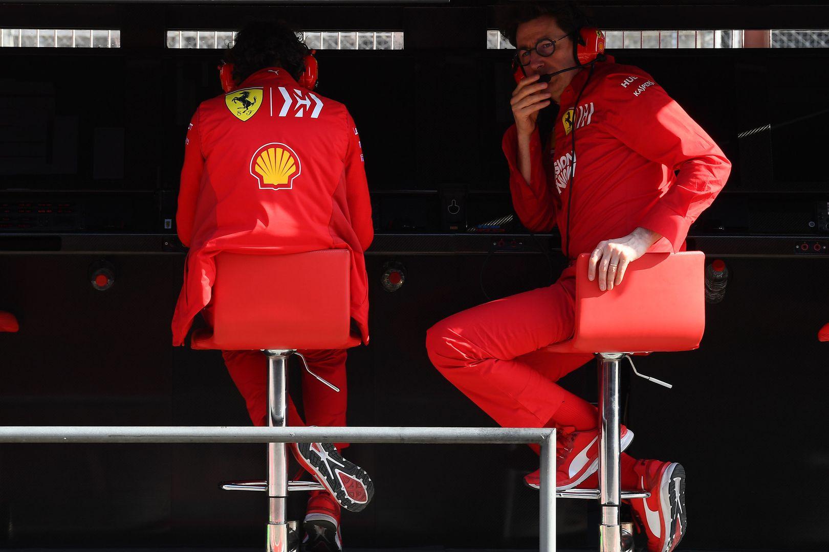Due membri della Scuderia Ferrari in tuta rossa sono seduti al muretto box, uno di loro è Mattia Binotto con cuffie e microfono, intento a comunicare via radio. Sullo sfondo si notano monitor ed il tipico allestimento da gara di Formula 1.