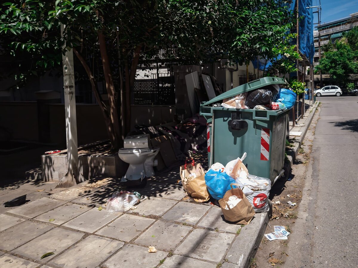 Cassonetto dell’immondizia stracolmo con sacchi e rifiuti abbandonati sul marciapiede, incluso un vecchio water.