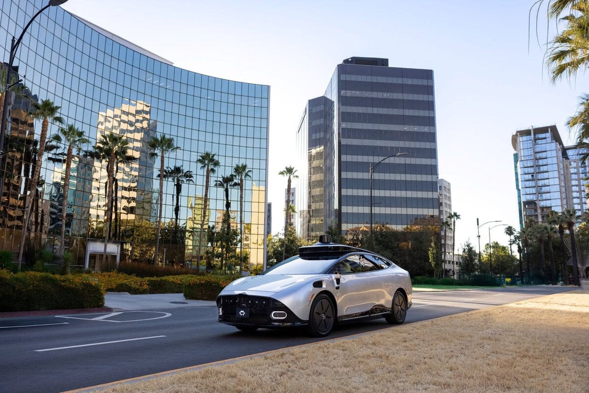 Auto a guida autonoma Tensor Robocar di colore argento che percorre una strada urbana con palme e grattacieli riflettenti.