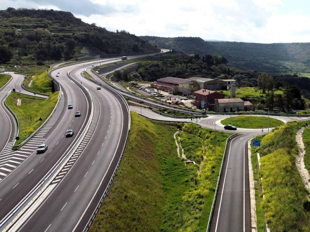 Tratto di autostrada con più corsie e svincolo immerso in un paesaggio collinare verde.