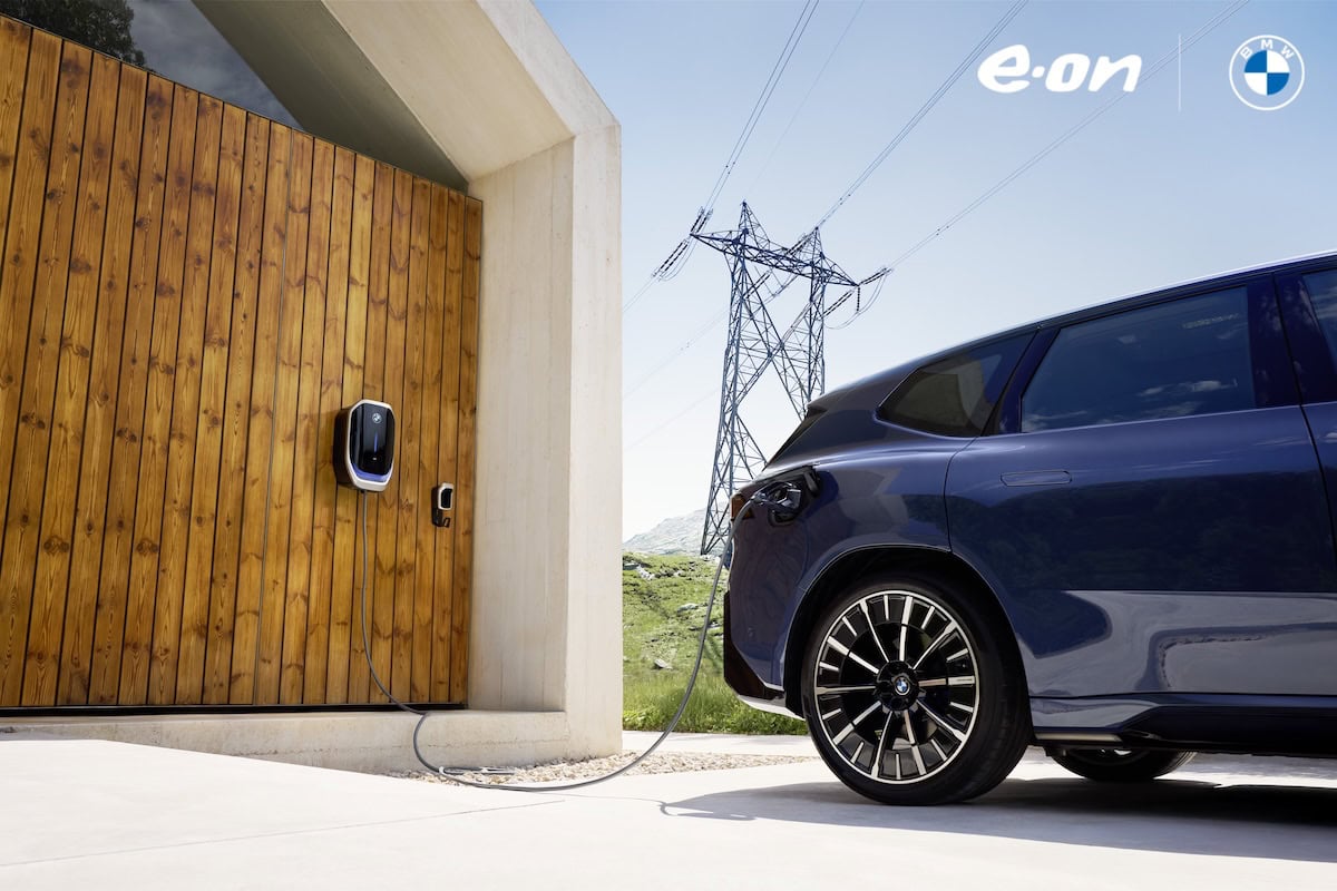 Auto elettrica BMW collegata a una wallbox E.ON installata su parete in legno, con tralicci elettrici sullo sfondo in un contesto montano soleggiato.