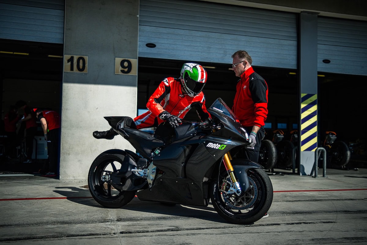 Pilota in tuta rossa sale sulla Ducati V21L elettrica nera da competizione MotoE 2025, assistito da un tecnico Ducati davanti ai box del circuito.