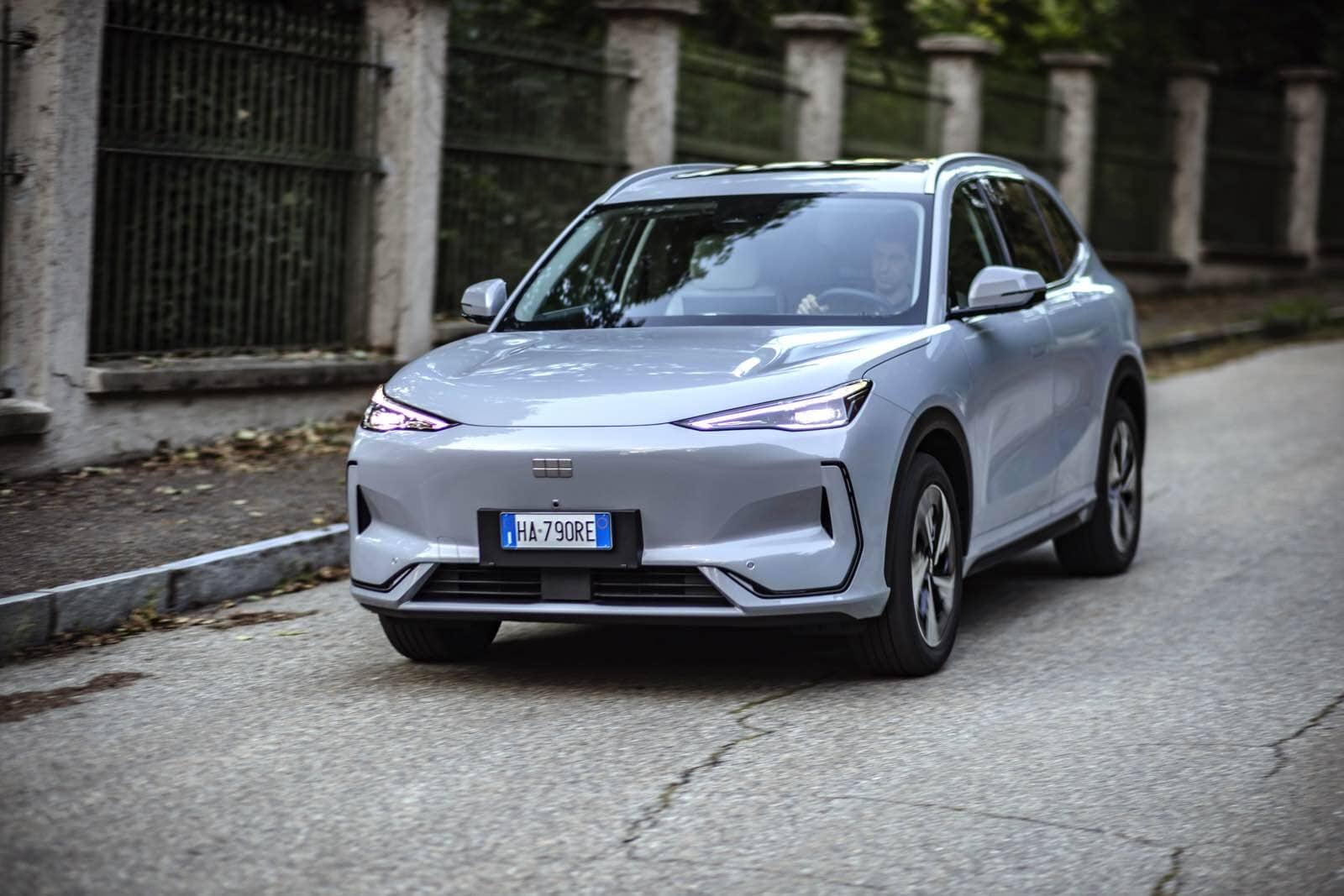 Auto elettrica Geely EX5 in movimento lungo strada urbana, design aerodinamico, tecnologia avanzata, veicolo sostenibile, auto 100% elettrica per città.