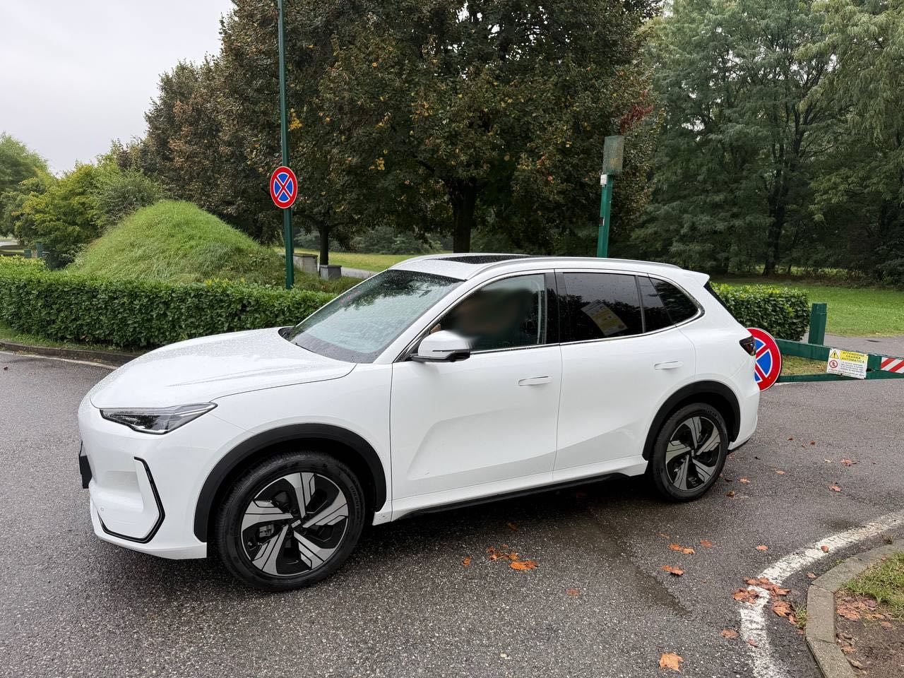 SUV elettrico Geely EX5 bianco visto lateralmente su una strada bagnata, in un contesto urbano con alberi e segnaletica stradale.