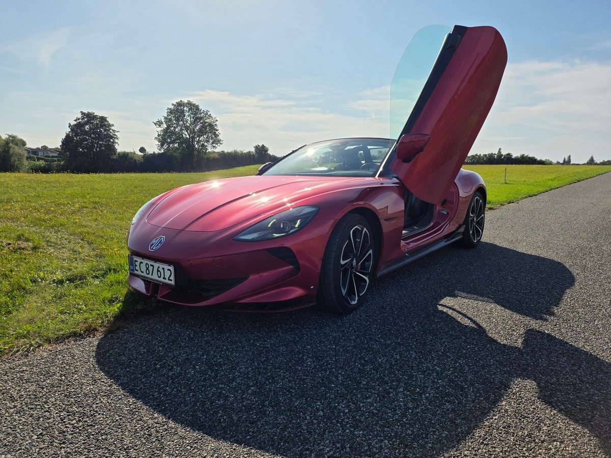 Convertibile sportiva rossa con porte verticali aperte, in posizione laterale su strada asfaltata in campagna.