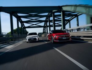 Due modelli di Lancia Ypsilon ibrida, una bianca e una rossa, corrono affiancate su un ponte moderno in acciaio durante una prova su strada. L’immagine mette in risalto il design sportivo e lo stile dinamico della nuova city car ibrida.