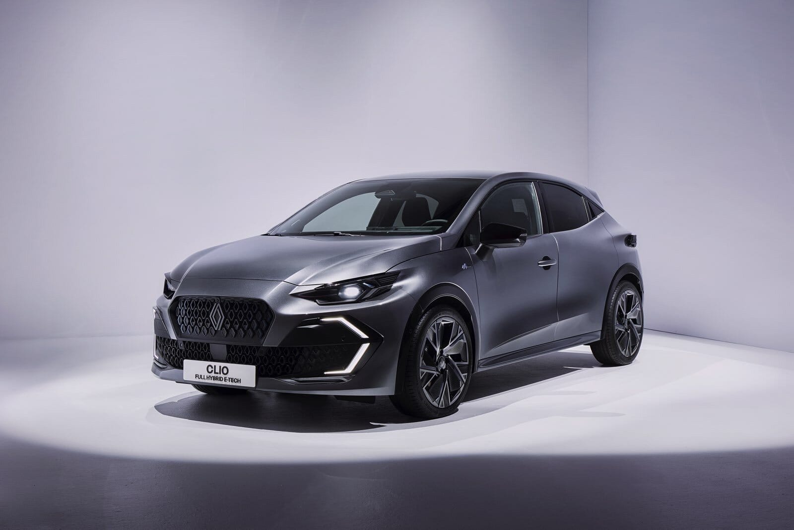 Renault Clio E-Tech fotografata da un angolo tre quarti anteriore sinistro, con fari accesi e dettagli sportivi in un set fotografico bianco.