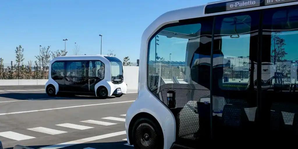 Due Toyota e-Palette a guida autonoma si incrociano su una strada cittadina, con carrozzeria bianca e nera e design futuristico simmetrico.