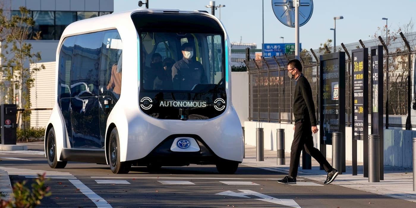 Toyota e-Palette autonomo in movimento in area urbana, con passeggeri a bordo e scritta "AUTONOMOUS" sul display frontale, mentre un pedone attraversa sulle strisce.