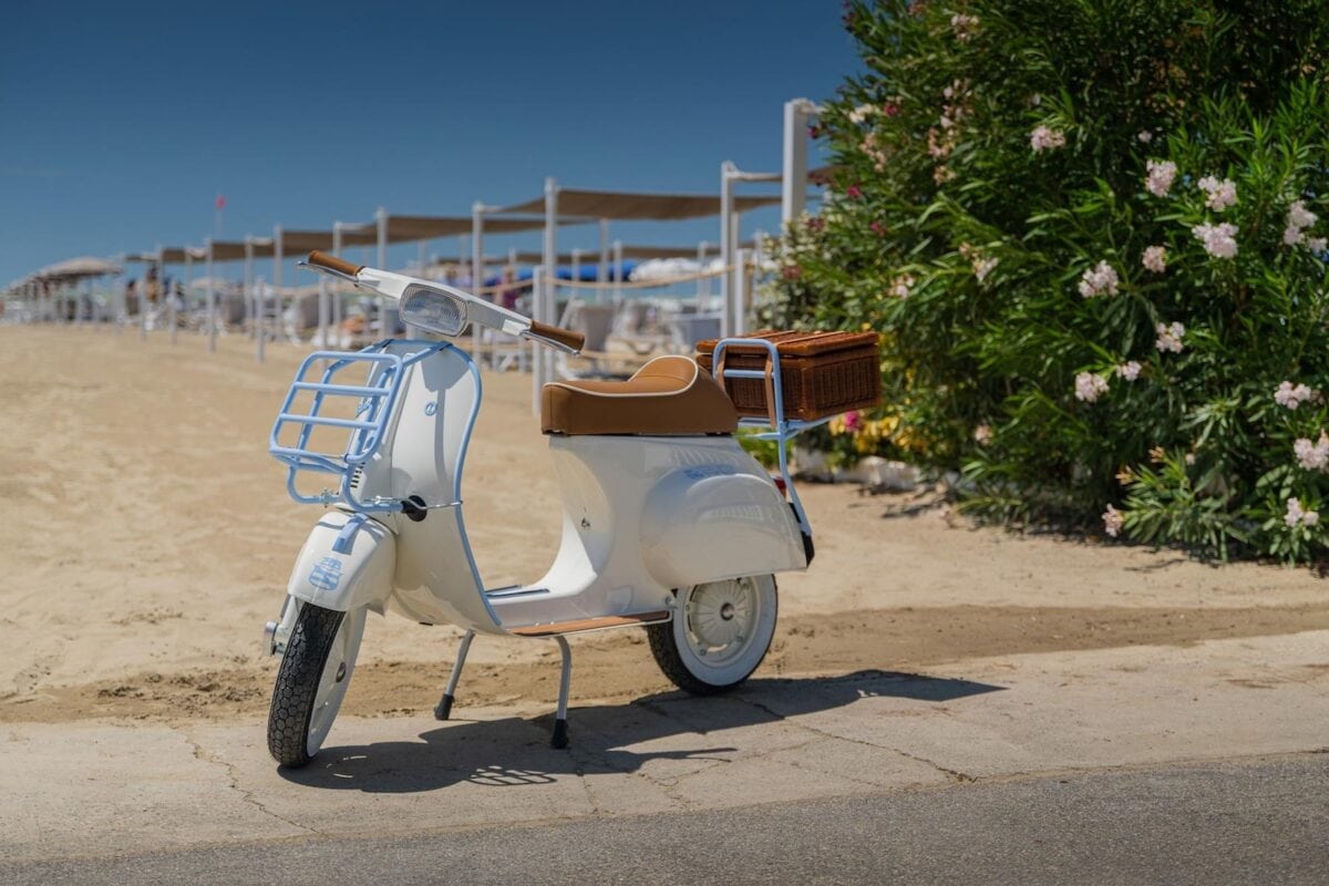 Una Vespa Special-e Garage Italia dallo stile vintage parcheggiata su una passeggiata sabbiosa vicino alla spiaggia, con cestino in vimini sul portapacchi posteriore e vegetazione fiorita sullo sfondo.