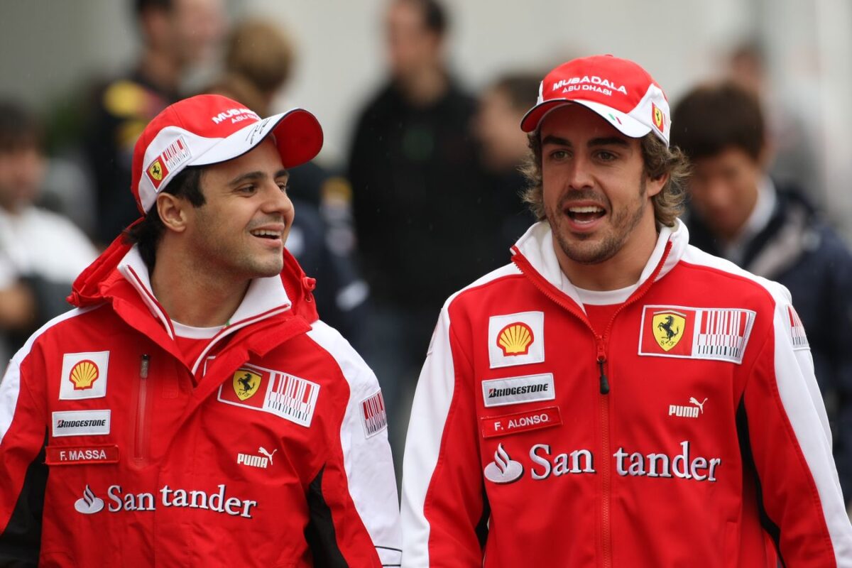 I piloti Ferrari Felipe Massa e Fernando Alonso sorridenti nei box durante un weekend di gara, entrambi in divisa rossa ufficiale con cappellino e loghi degli sponsor.