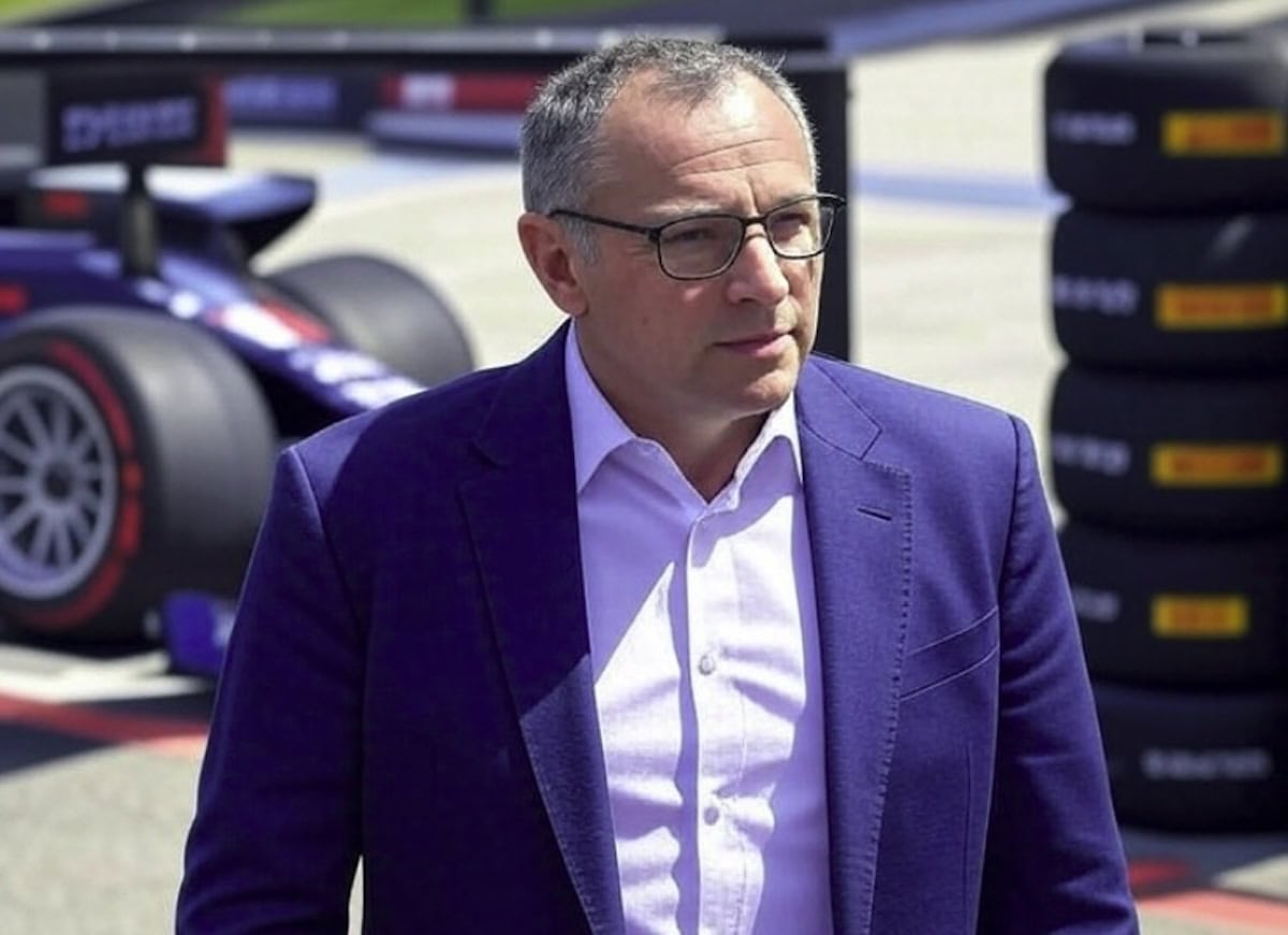 Stefano Domenicali, presidente della Formula 1, in abito blu e camicia bianca, cammina nel paddock con gomme Pirelli e una monoposto sullo sfondo.