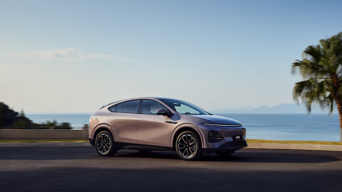 SUV coupé elettrico XPeng G6 in movimento su una strada costiera con vista sul mare, palme e montagne all’orizzonte, in una scena dal clima esotico.