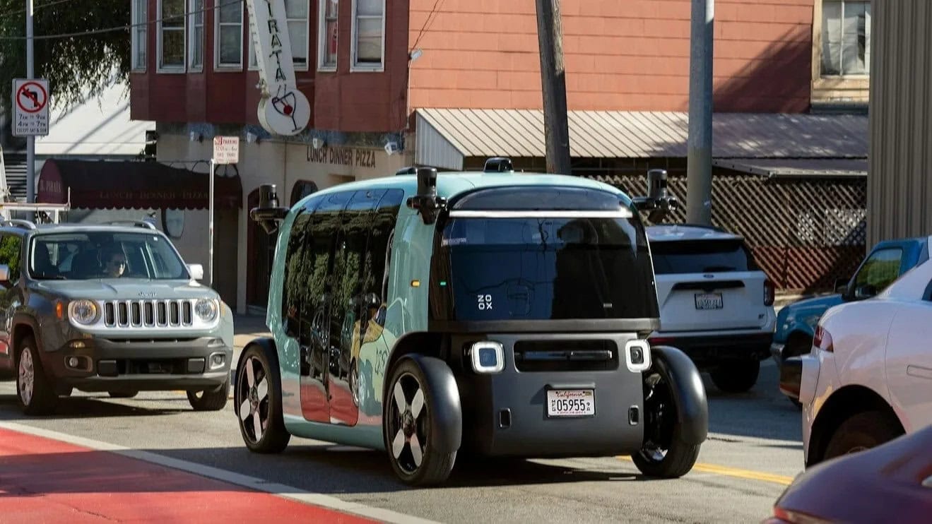 Robotaxi autonomo Zoox in marcia in un centro urbano, circondato da altre auto, con negozi e insegne visibili sullo sfondo.