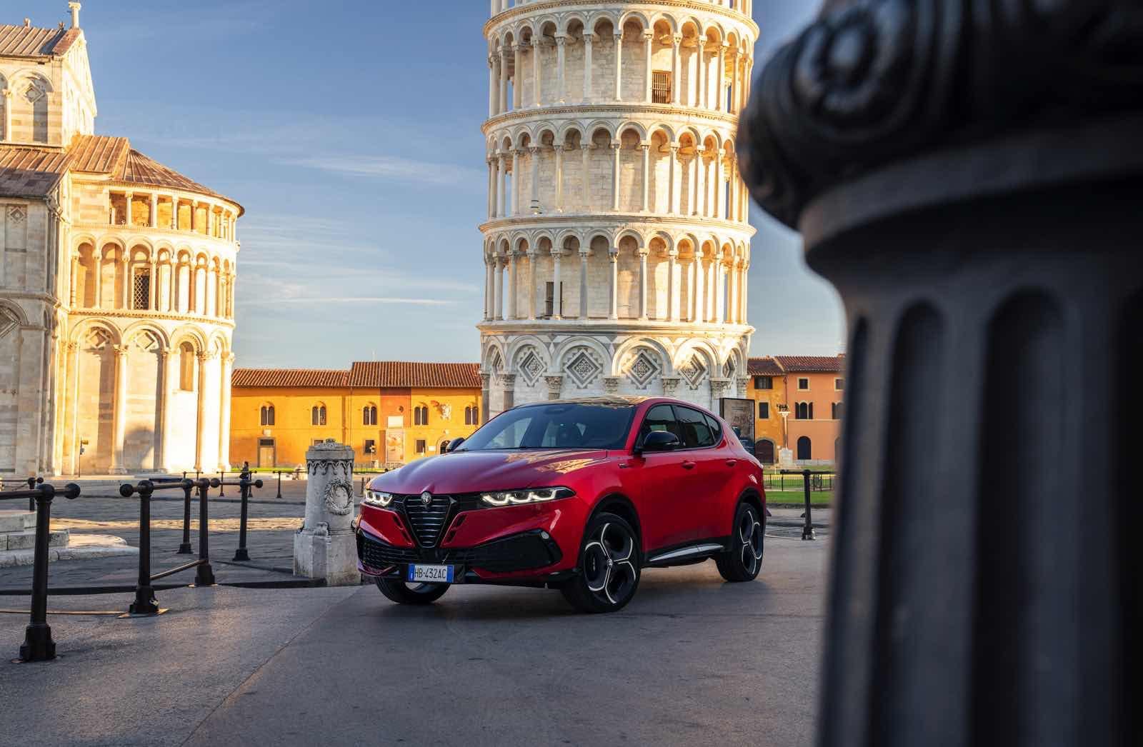 Alfa Romeo Tonale rossa in piazza con Torre Pendente di Pisa sullo sfondo, auto SUV di lusso, design elegante e prestazioni elevate, occasione di guida in Italia.