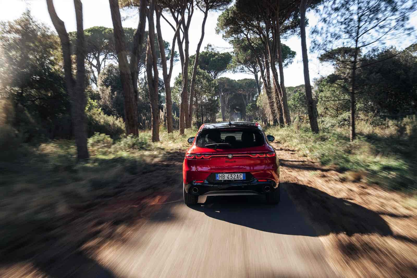 Auto sportiva Alfa Romeo Tonale MY 2026 in corsa su strada sterrata nel bosco, immagine dinamica del veicolo rosso in movimento tra gli alberi, test drive su percorso off-road, auto compatta e potente all'aperto.