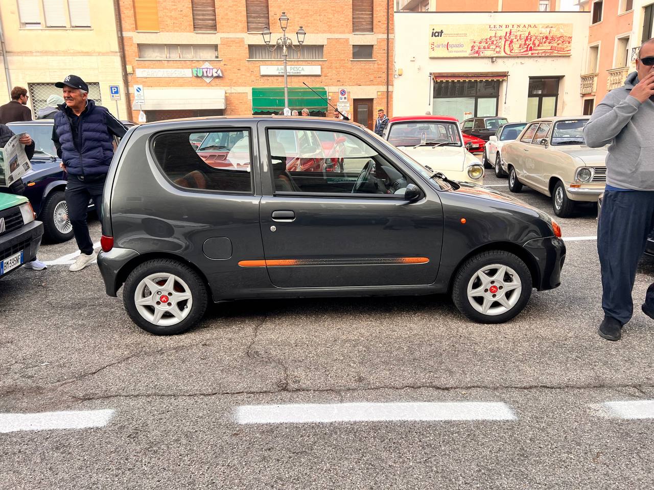 Fiat Seicento nero parcheggiato in strada con persone che lo ammirano durante un raduno di auto storiche.