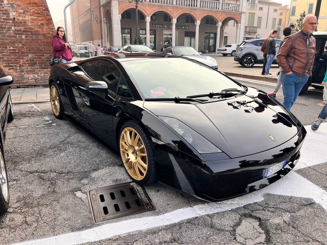 Lamborghini Gallardo nera con cerchi d'oro parcheggiata in strada in un centro urbano.