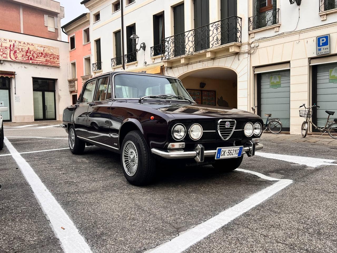 Alfa Romeo GTV degli anni '70 parcheggiato in una zona urbana con edifici storici sullo sfondo.